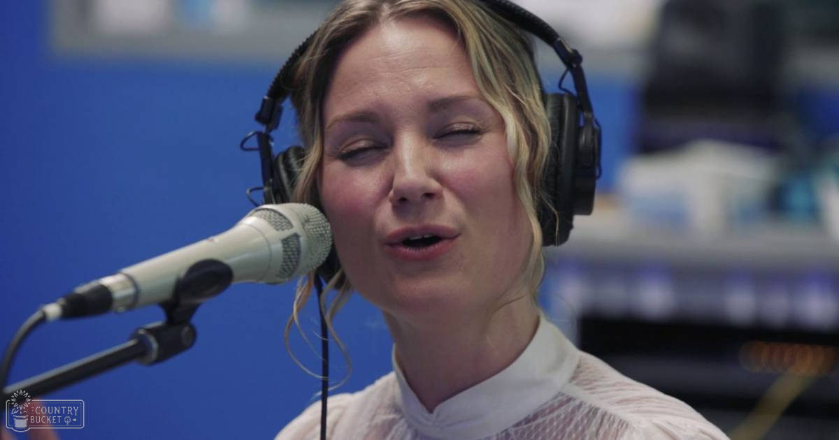 A young adult female in a white, textured blouse with short sleeves and large over-ear headphones is speaking into a microphone within an indoor recording studio setting.