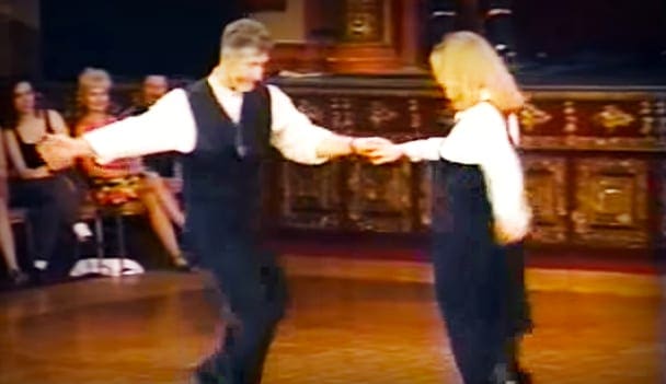 A couple dancing indoors; man in dark vest over a white shirt with collar and tie, woman in similar attire, ornate furniture in the background.