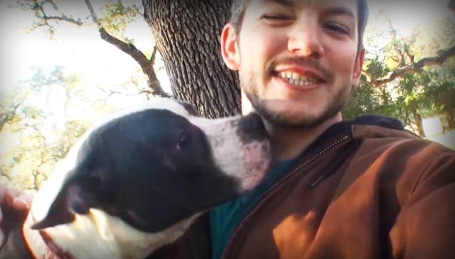 Young adult male wearing a brown jacket over a teal shirt, walking a dog in an outdoor setting with greenery and bright daylight.