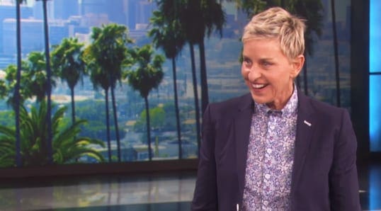 A middle-aged woman wearing a dark blue blazer and white blouse with purple and green floral patterns, standing indoors with a scenic cityscape and palm trees visible through large windows.