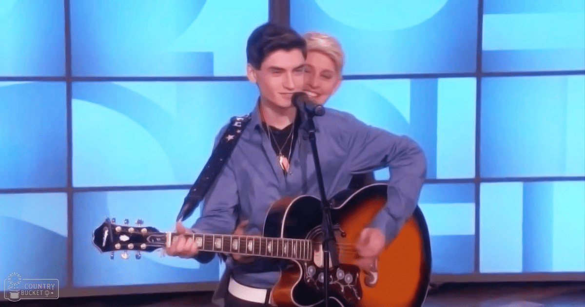 A young adult male wearing a loose-fitting blue button-up shirt performs on an acoustic guitar in a television studio setting with controlled artificial lighting.