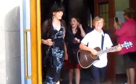 A young adult male plays guitar outdoors, wearing a white shirt with dark blue trim; an adult female holds a smartphone, dressed in a black floral dress. A child smiles in the foreground, wearing a dark graphic top and red shorts.