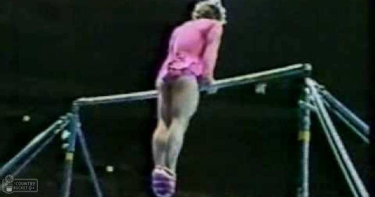 A young adult female performs gymnastics in an indoor arena wearing a form-fitting pink leotard with short sleeves, low-cut neckline, and athletic cut. The setting is a darkened indoor space typical of sports arenas or gymnastics facilities.