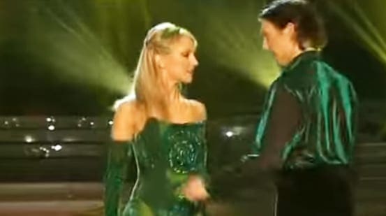 A woman wearing an elegant green off-the-shoulder dress with intricate detailing, styled in loose waves, stands on a stage illuminated by warm stage lights.