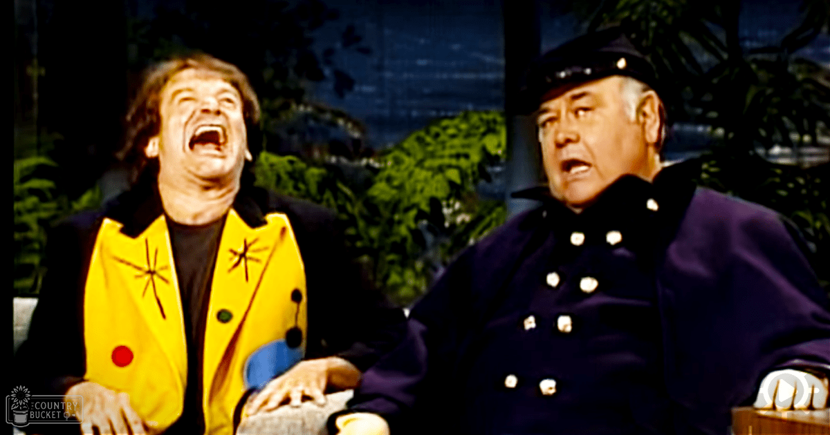 A comedic indoor studio scene with two middle-aged men, one in a bright yellow jacket and the other in a dark blue military-style coat. The setting includes greenery and artificial lighting.
