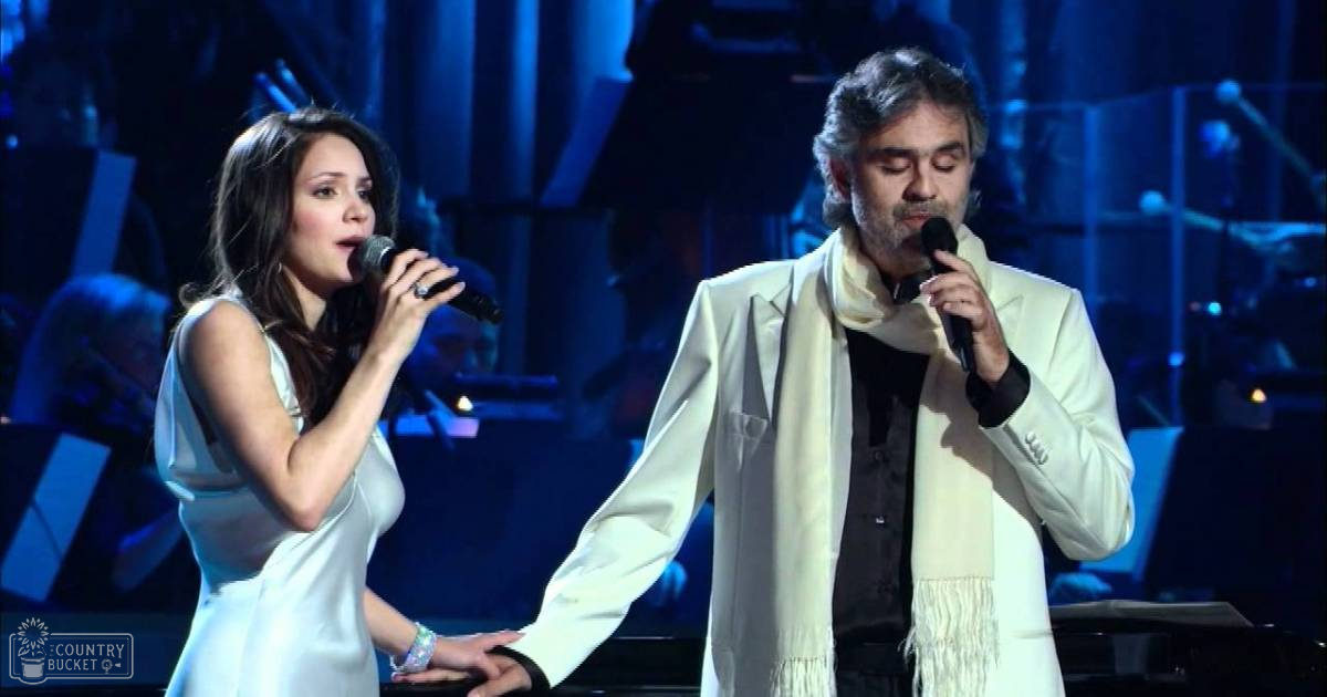 A young adult female singer dressed in a sleeveless white dress performs on stage alongside a middle-aged male conductor wearing a cream-colored suit, under dramatic blue and white stage lighting at an indoor concert hall.