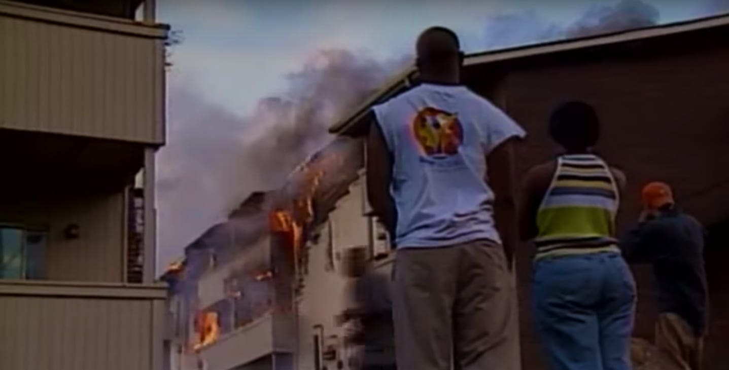 Adult male in light blue T-shirt and khaki pants observing fire with child wearing striped tank top and denim jeans; urban setting with buildings visible in background, late afternoon or early evening lighting conditions