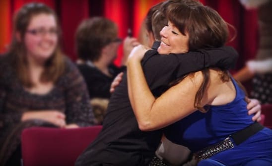 A woman with long brown hair in a dark patterned jersey top, an adult male wearing glasses and a fitted dark shirt, and another woman in a blue sleeveless top hugging someone. They are seated on red chairs indoors at what appears to be a talk show or event.