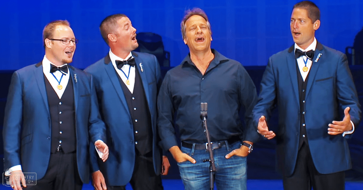 A group of adult males performing on a stage with blue lighting, wearing formal attire including suits and bow ties.