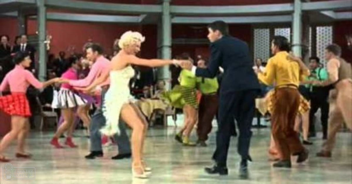 A dance scene with a group of young adults in colorful outfits dancing indoors under artificial light.