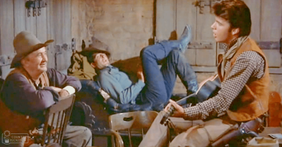 An older adult male wearing a dark hat, long-sleeved shirt with rolled-up sleeves, and light-colored pants sits among younger adults in a rustic cabin setting. The scene includes wooden furniture, hanging tools, and various mid-20th-century clothing items.