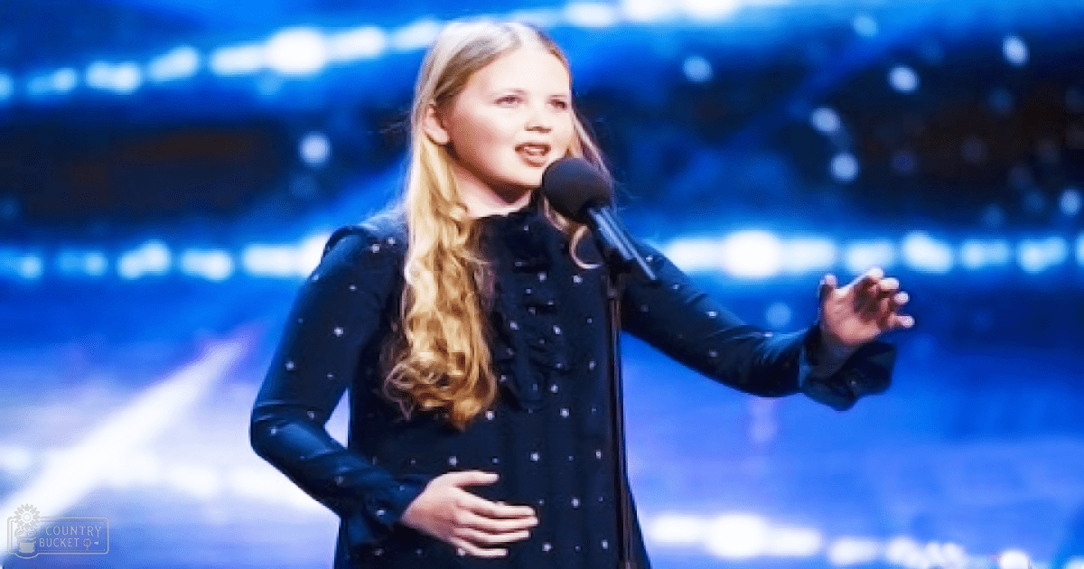 A young adult female sings on stage at an indoor performance venue with a blue-lit backdrop, wearing a dark blue or black button-up shirt with small white dots.