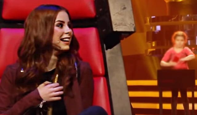 A young adult female participant in a singing competition, wearing a dark jacket with red leather chairs and stage lighting in the background.