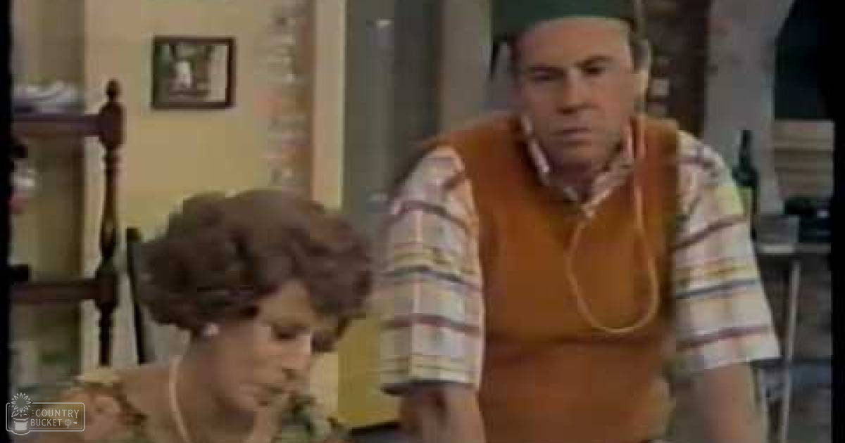 An older adult male in a home setting with children, light-colored top, striped shirt and orange vest, daytime indoor scene with staircase and furniture.