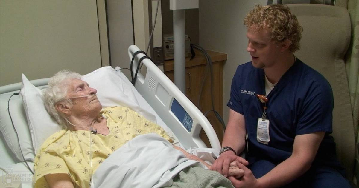 Elderly female in a hospital room with yellow floral-patterned gown, young male doctor in dark blue scrubs, modern medical equipment, indoor setting