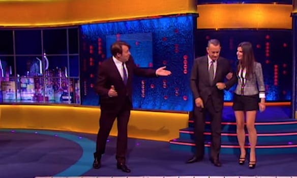 A talk show host and two guests in a modern television studio with abstract blue and yellow lighting.