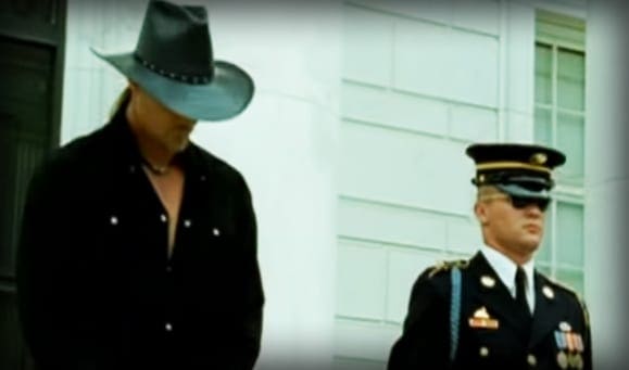 A man in a cowboy hat and another person in a military uniform stand outside an institutional building with white walls during daytime.