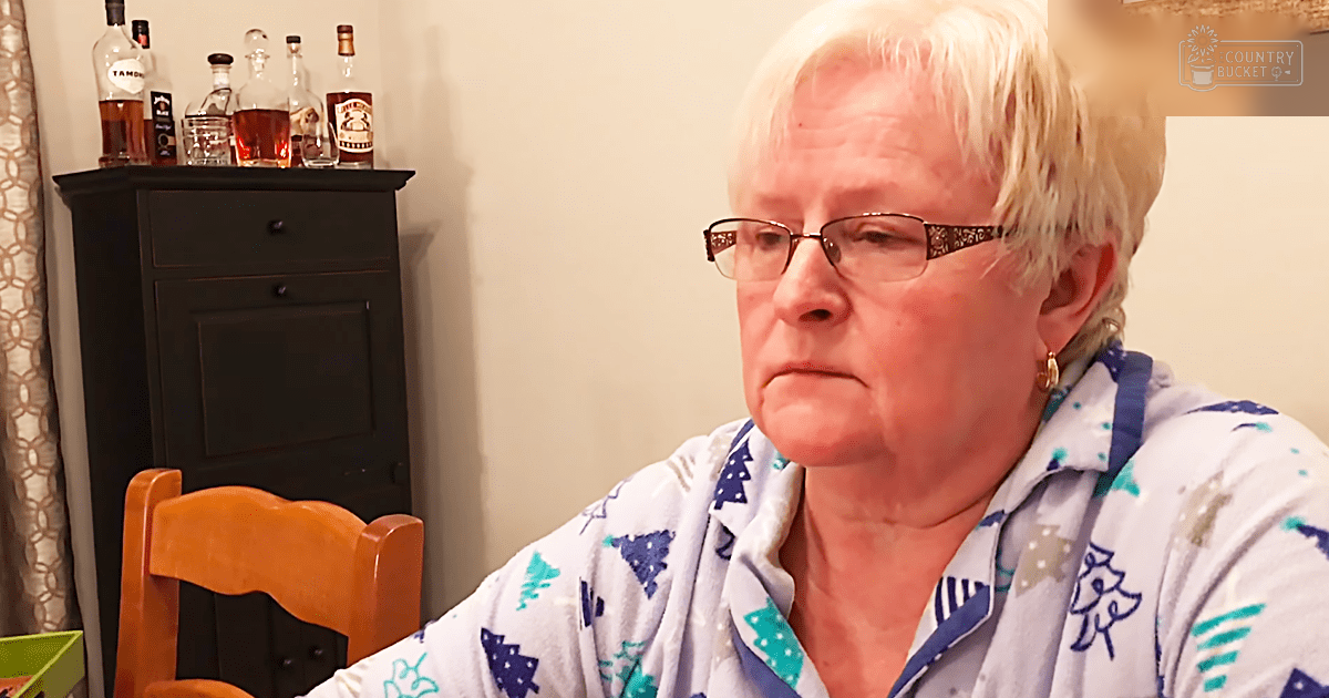 An older adult female in a home setting, wearing patterned Christmas-themed pajamas with long sleeves. The scene is indoors, possibly during winter.