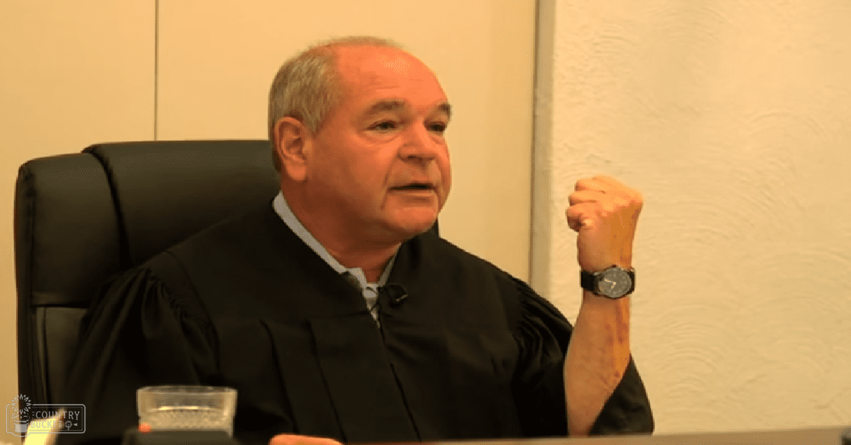 A middle-aged adult male in a black judicial robe sits at a desk, surrounded by indoor furniture and artificial lighting.