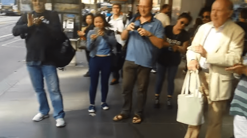 A group of pedestrians, including an adult male in jeans and a dark jacket holding a camera, and various others, are captured mid-movement on what appears to be an urban sidewalk or street. The scene is set during the daytime with clear daylight illuminating the subjects.