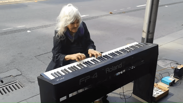 An older adult female plays a keyboard in an outdoor public space with overcast lighting, suggesting late fall or early spring.