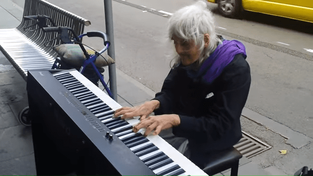 An older adult female plays a Yamaha keyboard in an urban setting with a blue bicycle and yellow vehicle nearby, under natural daylight.