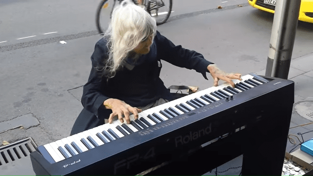 An older adult male singer plays a Roland FP-4 keyboard on an urban sidewalk, with scattered coins and a bicycle wheel nearby.