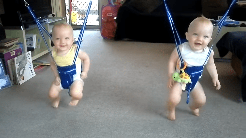 Two infants in a carpeted room with blue jumpers, furniture, books, and natural light.