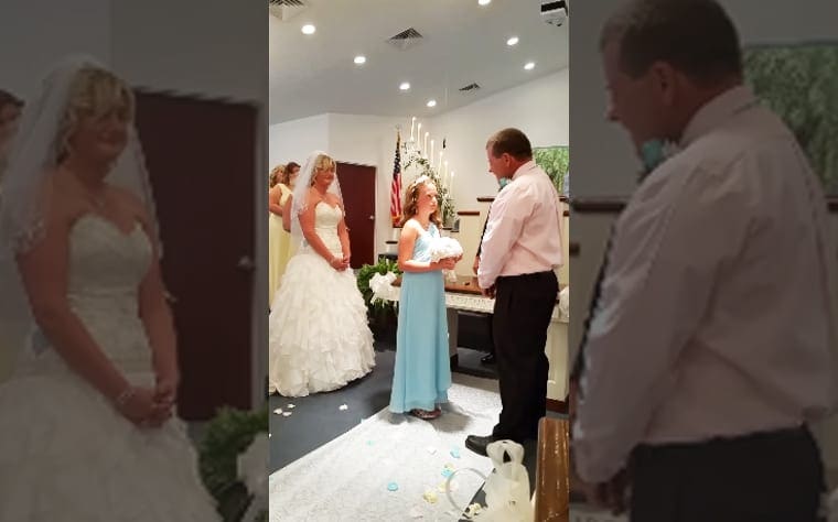 An adult female bride in a white strapless wedding gown with ruffled layers and a veil stands at her wedding ceremony, accompanied by a young girl holding flowers on the aisle. The setting is indoors, decorated with white flowers and petals, suggesting it's likely spring or summer.