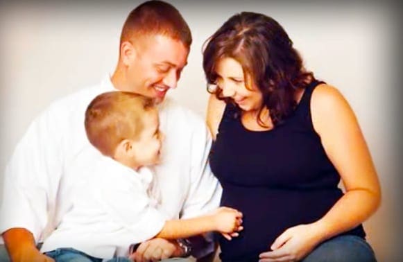 A family scene indoors with an adult male, a toddler, and an adult female against a plain white wall under soft artificial lighting.