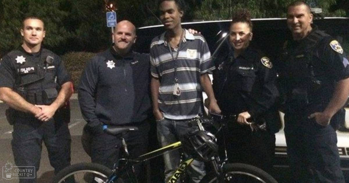 A group of law enforcement officers and a community member pose outdoors near what seems like a police station with a bicycle in front.