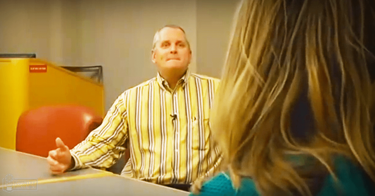 A middle-aged man wearing a yellow striped button-up shirt sits in an office setting with a young adult female in a teal knitted sweater nearby. The scene includes red furniture and some yellow equipment, suggesting a casual or business-casual environment.