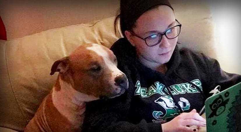 A young adult female wearing a dark-colored Seahawks hoodie, reading while holding a tablet with a green cartoon character cover, accompanied by an American Pit Bull Terrier or similar breed dog in a beige living room setting under indoor lighting.