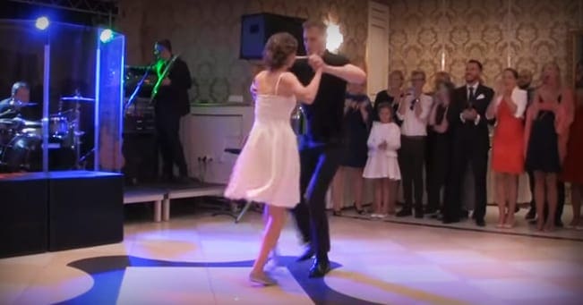 A young couple dancing at an indoor banquet hall during a formal event. The female dancer wears a white dress with thin straps and black shoes, while the male dancer is in dark pants and shirt. An audience of adults dressed formally claps along.