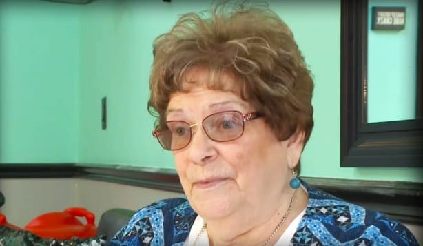 An older adult female wearing glasses, earrings, and a patterned blue and white top with a necklace indoors in front of green walls under natural light.
