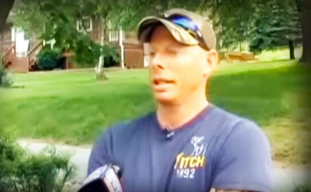 A young adult male in a relaxed fit crew-neck T-shirt with short sleeves, wearing a baseball cap and sunglasses, holding what appears to be a microphone or recording device, standing outdoors on green grass with trees and part of a house visible in the background.