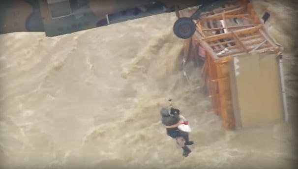Rescue operation in progress at an urban flood scene; adult males in waterproof suits assist amidst murky, turbulent floodwaters under overcast skies.