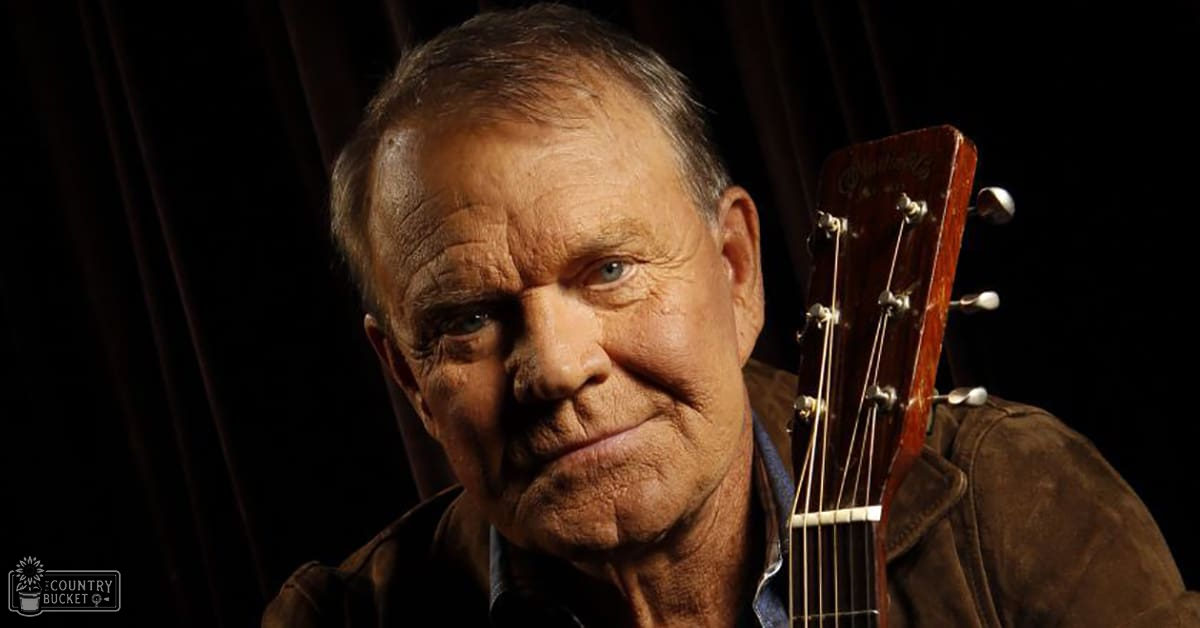 An older adult male in a dimly lit indoor setting, wearing a brown jacket with visible stitching and a collar underneath. A guitar with a red headstock is partially visible on the right side of the frame.