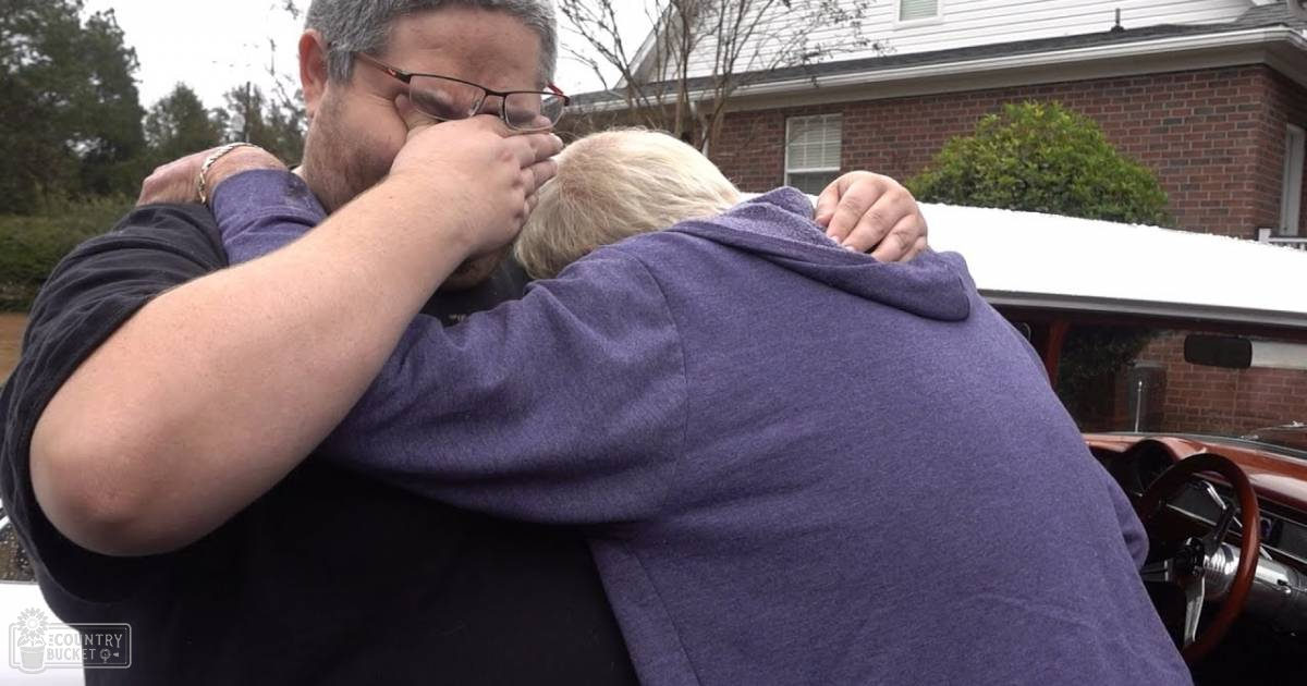 An adult male singer hugs an older adult woman outside a house with snow on the ground, wearing glasses and a watch. The woman wears a purple hoodie.