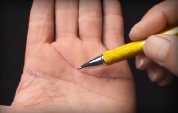 A hand holding a yellow pen with black tip, making marks on another palm in an indoor setting with dark and out-of-focus background