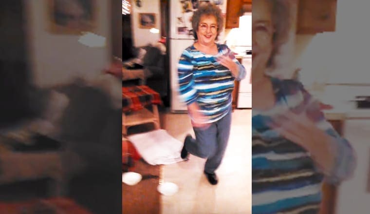 An older adult female in a home setting, wearing a blue and white striped long-sleeve shirt with horizontal stripes, blue jeans, black shoes. The scene includes kitchen cabinets, appliances, and various items on a table.