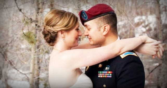 A couple in a forest setting; the woman wears a white wedding dress and earrings, while the man is dressed in military uniform with medals and a beret.