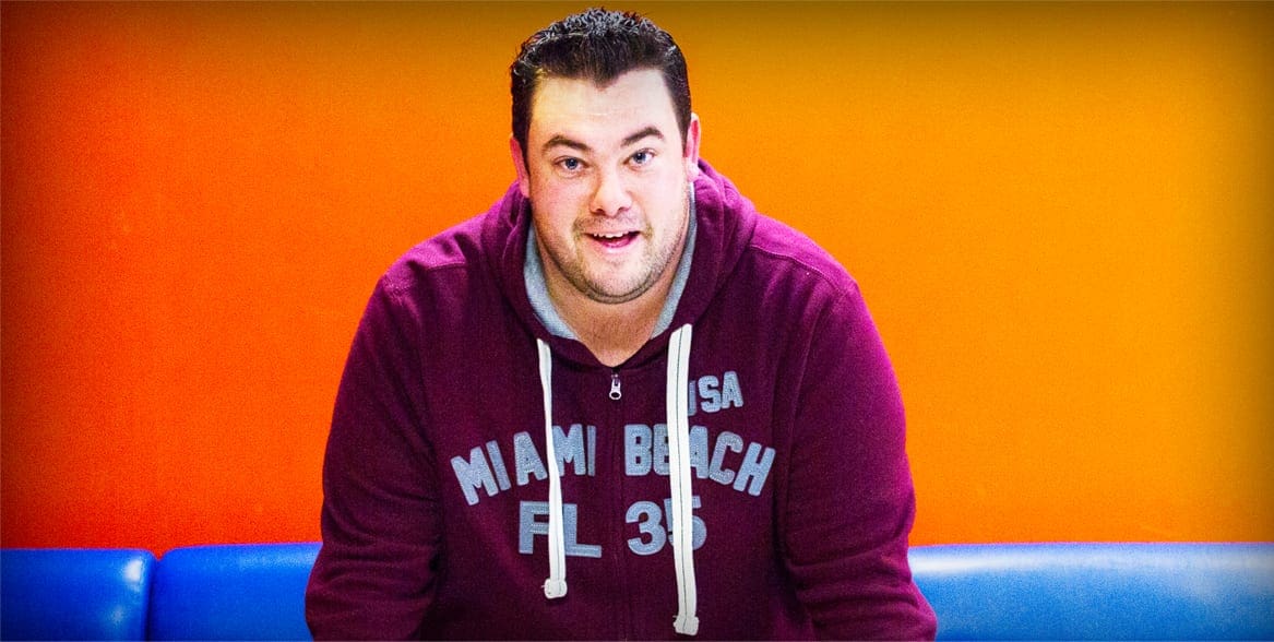 An adult male in a casual maroon hoodie with 'Miami Beach FL 35' printed across the front, sitting on a blue couch against an orange wall. The setting appears to be indoors during daytime.
