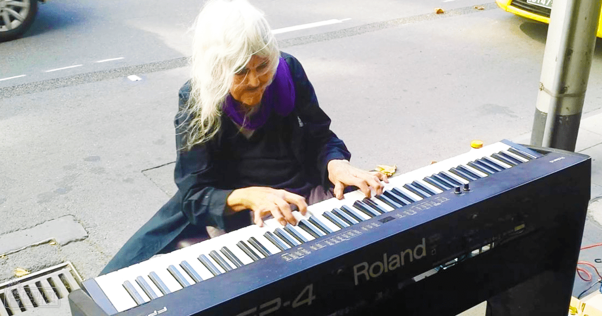 An older adult female wearing a dark blue jacket and purple scarf plays a Roland TP-4 keyboard in an outdoor urban setting with wet pavement, possibly spring or summer.