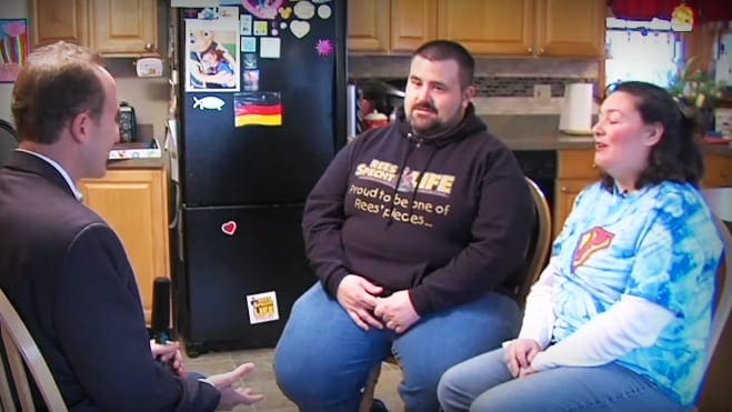 An adult male in a kitchen setting with two other middle-aged individuals, discussing or interviewing each other against a backdrop of wooden cabinets and various items on the counter, including magnets and papers.
