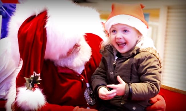 A toddler girl in a brown jacket interacts with an older adult male Santa Claus wearing a red suit and white fur trim, set indoors with light-colored walls and blinds.