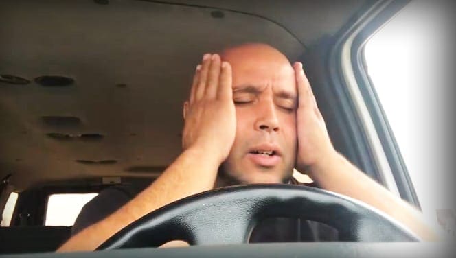 A middle-aged to older adult male wearing a dark-colored t-shirt with a simple design sits inside a car, showcasing part of the steering wheel and dashboard.
