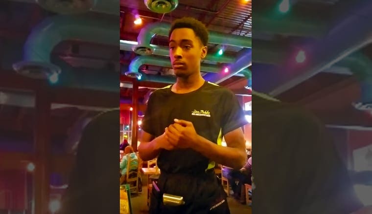 A young adult male wearing a 'Dan Paddle' t-shirt sits in a casual indoor setting with decorative lighting and furniture suggesting a restaurant or café environment.