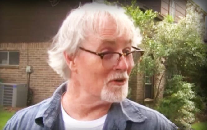 Older adult male in a light blue button-up shirt over a white t-shirt, standing outdoors with trees and greenery surrounding a brick house on a sunny day.