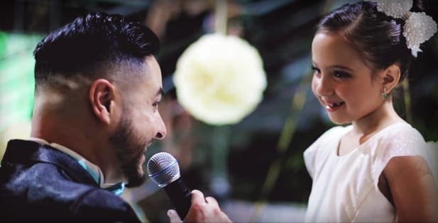 An adult male singer speaks into a microphone with a young girl in a white dress beside him, set outdoors amidst green foliage under soft daytime light.